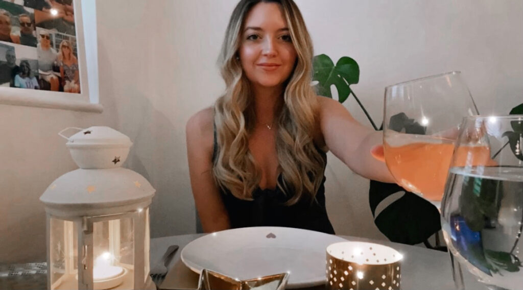 Rosie is sitting at a grey dining table holding out a glass of white wine to cheers. On the table are candles and a white plate. Behind her is a monstera plant.