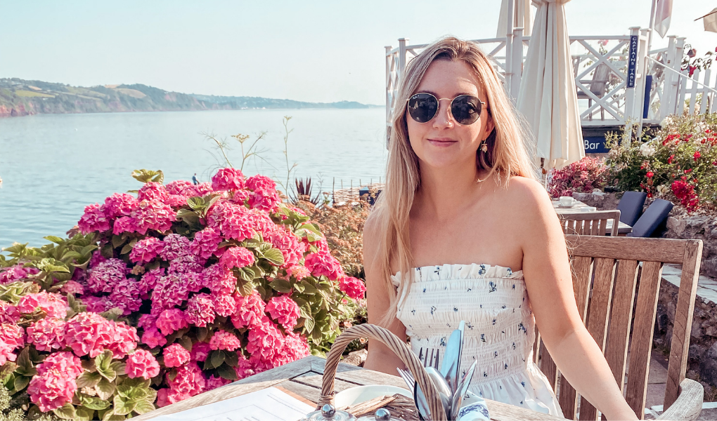 Rosie sat at a table, wearing a white dress and round sunglasses, beside pink flowers infront of a sea view. 